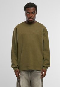 Oliņu zaļas teksturētas sporta krekls ar brīvu piegriezumu, apaļu kakla izgriezumu un garām piedurknēm. Iekļauj diskrētu zaļu izšūtu logotipu.