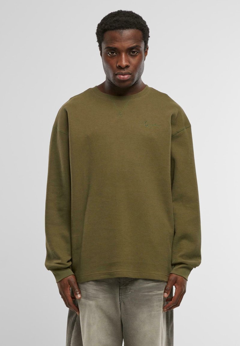 Oliņu zaļas teksturētas sporta krekls ar brīvu piegriezumu, apaļu kakla izgriezumu un garām piedurknēm. Iekļauj diskrētu zaļu izšūtu logotipu.