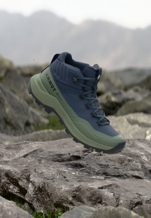 Botte de randonnée bleue et verte avec une semelle rugueuse flottant au-dessus d'un terrain rocheux dans un cadre extérieur montagneux.