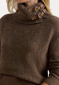 Hnedý pletený sveter s vysokým rebrovaným golierom, ktorý má dva kožené popruhy a kovové pracky na zapínanie. Textúrovaná, teplá látka.