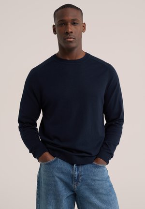 Mann trägt einen marineblauen Pullover mit langen Ärmeln und hellblaue Jeans und steht mit den Händen in den Taschen vor einem einfarbigen Hintergrund.