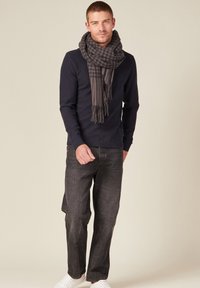 Camicia a maniche lunghe blu navy, jeans grigi scuri, sneaker bianche e una sciarpa scozzese nera e grigia con frange, indossati in modo casual.