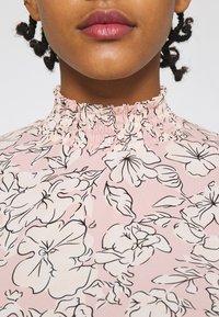 Rosa blus med blommor och smockad halsringning. Har svarta konturer av blommor och blad mot en mjuk rosa bakgrund. Slät textur.