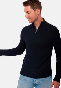 Pull à zip bleu marine avec texture côtelée et col montant. Présente une doublure jaune sous le col et un design ajusté.