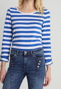 Blau- und weiß gestreiftes Langarmhemd, kombiniert mit dunklen Jeans aus Denim, die seitliche Metallknopf-Details aufweisen. Klassische Passform mit rundem Ausschnitt.