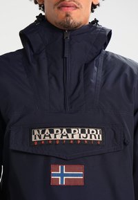 Anorak de color azul marino con cierre frontal, parche del logo que dice "Napapijri geographic" y acento de bandera bordado.