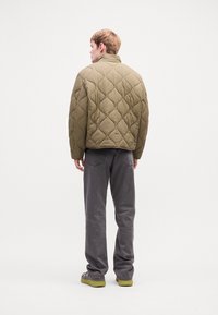 Persona de pie de espaldas, vistiendo una chaqueta acolchada verde olivo, pantalones vaqueros gris rectos y zapatos verde claro, sobre un fondo blanco.