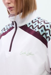 Λευκό πουλό βερ με φερμουάρ στο γιακά, με μαροον λεπτομέρειες και γεωμετρικό σχέδιο στον ώμο σε αποχρώσεις του aqua και καφέ. Διαθέτει κεντημένο λογότυπο.