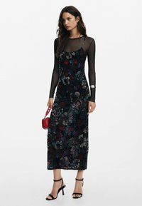Černé květinové šaty midi délky s průhledným topem s dlouhým rukávem, složitými vzory v červené, zelené a modré barvě, doplněné černými podpatky.