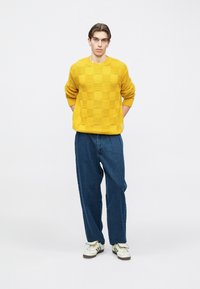 Mladý muž stojaci s rukami za chrbtom, oblečený v žltom kockovanom svetri, voľných modrých džínsoch a bielych teniskách na jednoduchom pozadí.