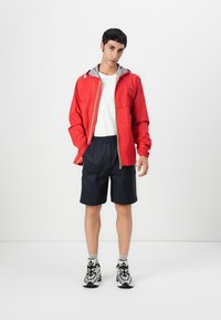 Rød vindjakke med grå hette, glidelås foran og elastiske mansjetter; paret med marineblå shorts og svart-hvite joggesko.