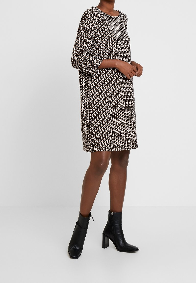 Kurzärmliges knielanges Kleid in Schwarz und Creme mit geometrischem Muster; lockere Passform; getragen mit schwarzen Ankle Boots mit Blockabsatz.