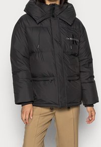 Svart puffjacka med stor krage, framtida dragkedja, dragsko-detalar och två sidfickor. Har subtilt varumärke på bröstet.