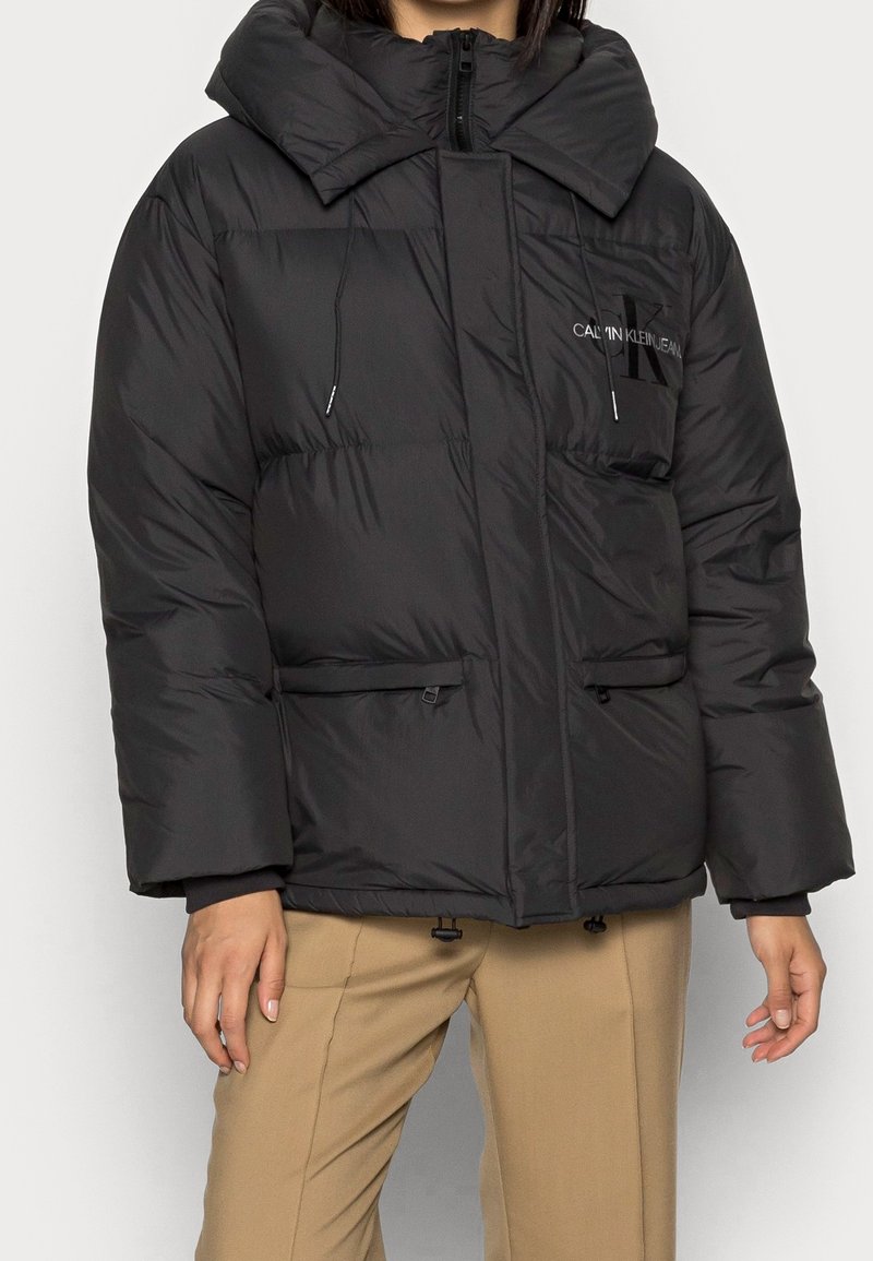 Svart puffjacka med stor krage, framtida dragkedja, dragsko-detalar och två sidfickor. Har subtilt varumärke på bröstet.