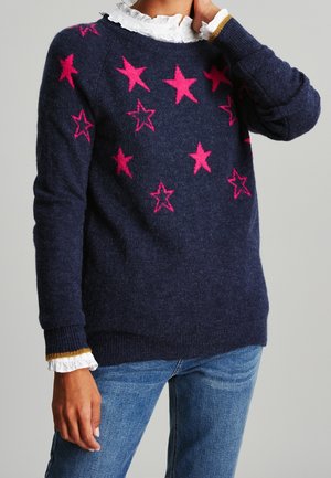 Personne portant un pull bleu marine avec des étoiles rose vif, un col et des poignets blancs à volants, associé à un jean bleu, debout devant un fond gris.