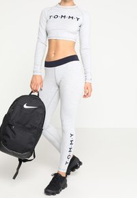 Grauer langärmeliger Cropped-Top mit dem Schriftzug "T.O.M.M.Y.", kombiniert mit passenden grauen Leggings, die seitlich Text zeigen, einem schwarzen Nike-Rucksack und schwarzen Sneakers.