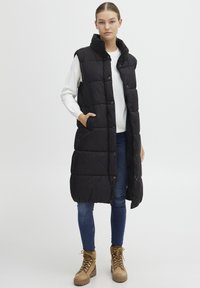 Schwarze Steppweste mit hohem Kragen, Druckknöpfen und seitlichen Taschen. Über einem weißen Pullover getragen und kombiniert mit blauen Jeans und beigen Stiefeln.