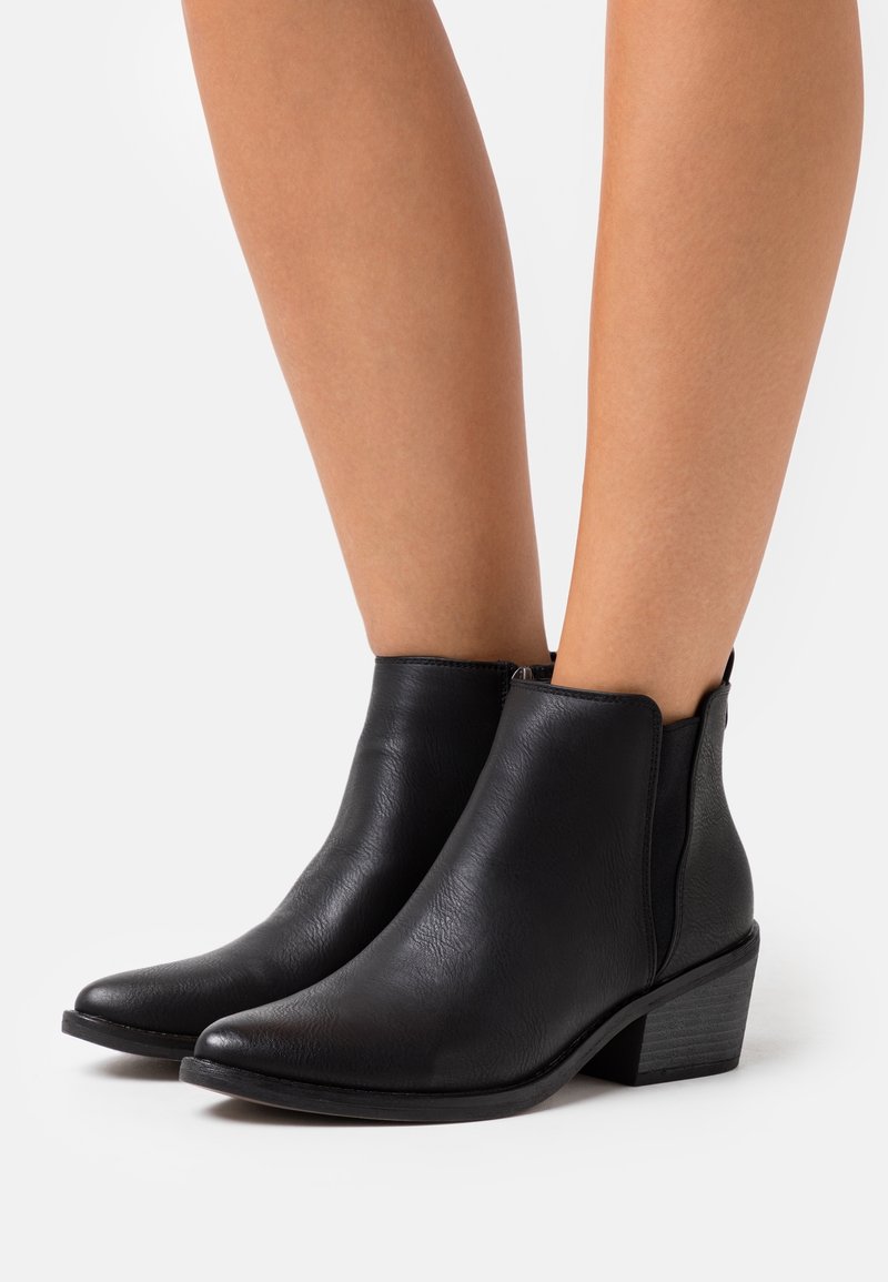 Bottes basses en cuir noir à bouts pointus et talons blocs portées sur des jambes nues sur fond blanc.