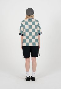 Person iført en grøn- og hvidternet kortærmet sweater, sorte shorts, hvide sokker, sorte sko og en grå flat cap, stående med ryggen til.