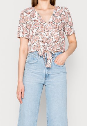 Femme portant un chemisier à manches courtes, à motif cachemire, noué devant, avec un jean taille haute bleu clair, une main dans la poche, fond neutre.