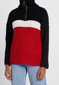 Tröja i stickat med hög krage; har svarta ärmar, röd kropp och vit rand över bröstet. Dragkedjesmycke vid halsringningen.