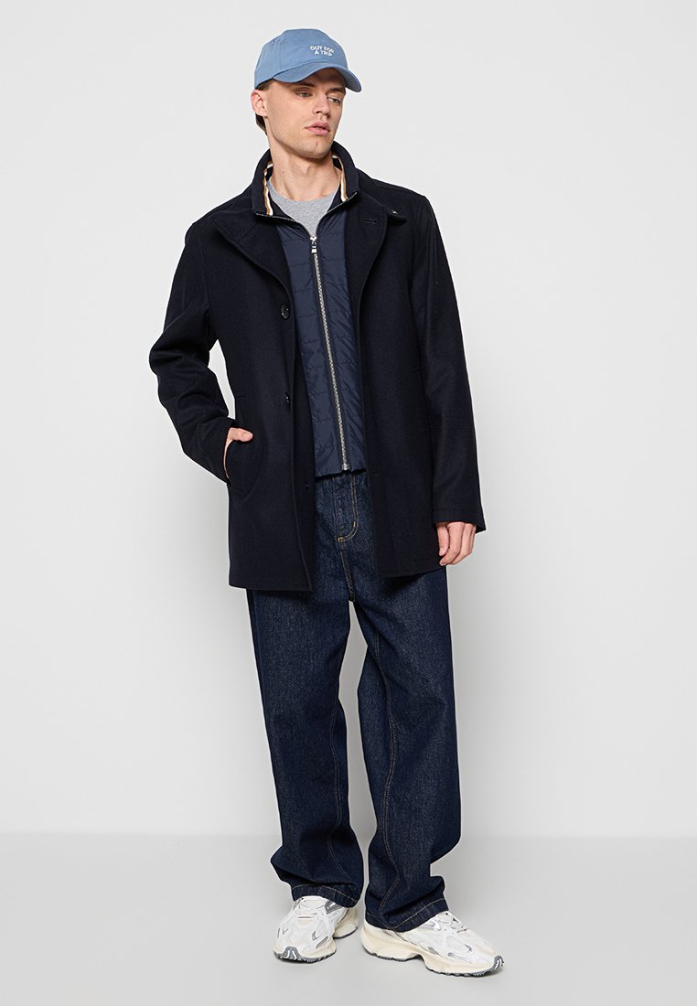 Cappotto di lana navy sopra una giacca trapuntata navy, abbinato a jeans scuri e sneakers bianche, completato da un cappellino azzurro chiaro.