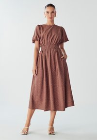 Robe midi marron à pois avec des manches courtes bouffantes et une taille élastique. Comprend des poches latérales et une silhouette fluide. Portée avec des sandales blanches.