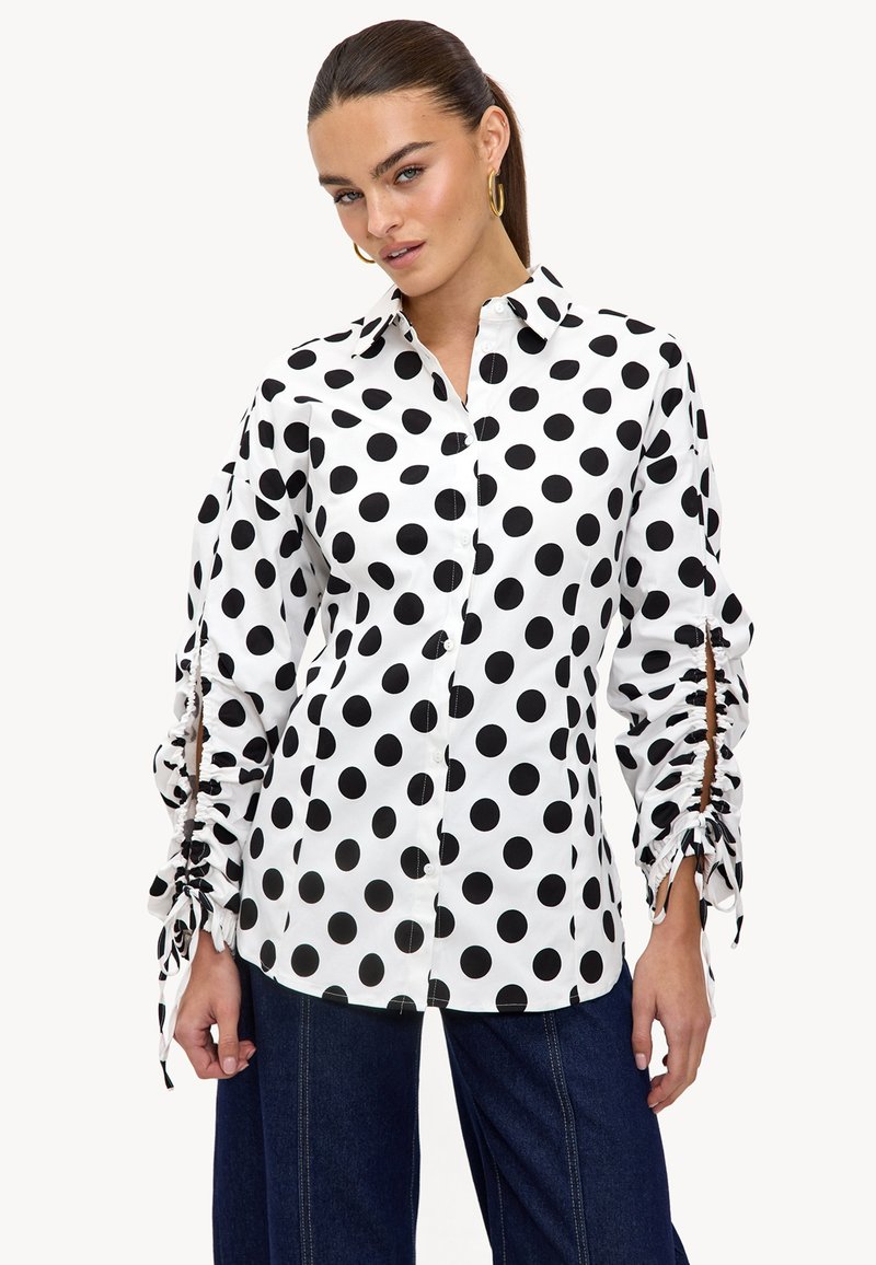 Chemise blanche à pois noirs, design à boutons, manches longues avec des accents froncés et des fentes, fabriquée en tissu léger.