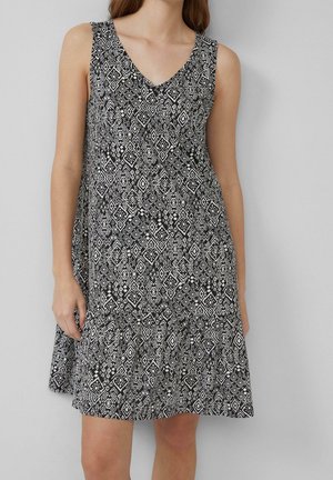 Černobílé geometricky vzorované šaty bez rukávů, sahající ke kolenům, s výstřihem do V a volánkovým lemem, které má na sobě stojící žena.