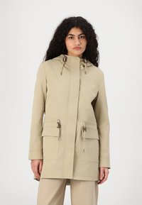 Cappotto beige con cappuccio e taglio dritto, caratterizzato da cordini e tasche frontali. Realizzato in un tessuto liscio e resistente.