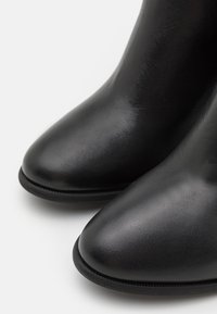 Botine negre din piele cu o textură netedă, vârf ascuțit și siluetă îngustă. Dispun de un detaliu subtil de cusătură pe marginea tălpii.