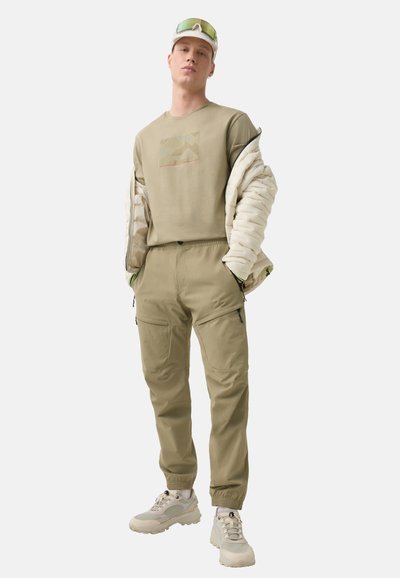 Khaki koszula z długim rękawem z grafiką gór, beżowa kurtka puchowa, pasujące spodnie cargo i jasne sneakersy z teksturowanym wzorem.