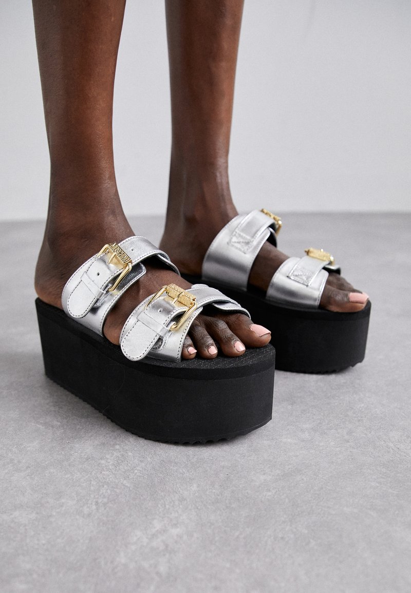 Zilveren platform sandalen met twee brede banden, gouden gespen en een gestructureerde zwarte rubberen zool. Het open teen ontwerp toont blote voeten.
