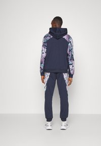 Marinblå träningsoverall med huva, zip-jacka med mönstrade ärmar och matchande avsmalnande byxor. Inkluderar vita accentband på sidorna.