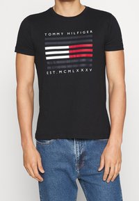 Černé bavlněné tričko s předním potiskem, který obsahuje horizontální pruhy v červené, bílé a námořnické modři, a text "TOMMY HILFIGER" nad tím.