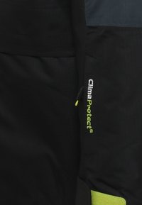 Un capo di abbigliamento esterno nero con un accento verde sulla tasca. Logo ricamato in bianco e verde, con una texture liscia e un design snellito.