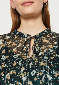 Tmavě zelená květinová blůza s plisé texturou, s detaily ve tvaru klíčové dírky a vázáním na krku. Zdobena béžovými a světle modrými květinami.