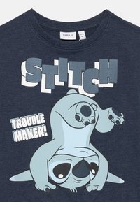 T-shirt in cotone blu navy con grafica "Stitch", illustrazione del personaggio sottosopra e il testo "TROUBLE MAKER!" in bianco bold.