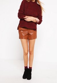 Rostfärgad strukturtröja med hög krage, kombinerad med bruna kortbyxor i konstläder och svarta ankelskor. Vinröda strumpor synliga ovanför skorna.