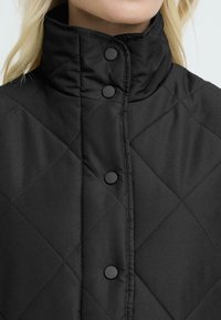 Sort quilted jakke med høj krave, knaplukninger og en glat tekstur. Tøjet ser let og vandafvisende ud.
