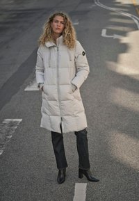 Abrigo largo acolchado de color beige claro, con capucha y cremallera. Se lleva sobre vaqueros oscuros y botines hasta el tobillo. Cuenta con dos bolsillos laterales y detalle de logo.