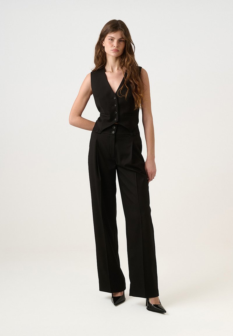 Juodas berankovis liemuo su priekiniais mygtukais, derinamas su aukšta juosmens plataus kirpimo kelnėmis. Lygi tekstūra ir pritaikytas siluetas. Aukšti kulniukai.