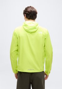 Άνδρας που φοράει φωτεινό κίτρινο-πράσινο αδιάβροχο μπουφάν με κουκούλα, στέκεται με την πλάτη προς το θεατή σε απλό λευκό φόντο.