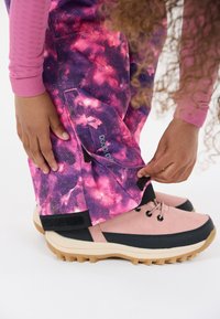 Pink and black hiking shoes with textured soles next to purple and pink galaxy-patterned pants. Pants feature velcro accents.