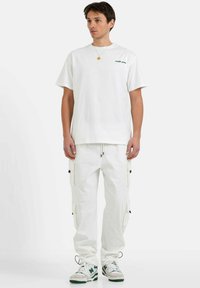 Witte katoenen t-shirt met korte mouwen en groene tekst op de borst, gecombineerd met witte cargobroek met zijzakken en een koord.
