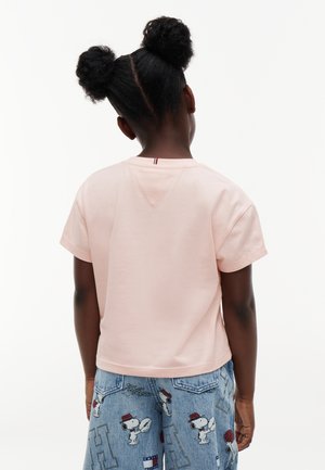 Barn med to puffede hestehaler iført en lyserød kortærmet skjorte og blå denimshorts med Snoopy tegneseriefigurer.