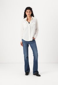 Chemise blanche à manches longues avec deux poches poitrine et fermeture à boutons, associée à un jean évasé bleu et des chaussures noires. Design simple et décontracté.