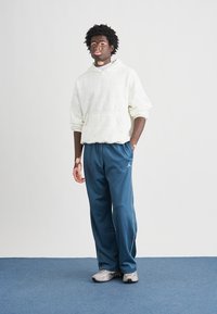 Sweat à capuche blanc avec logo texturé et poche kangourou, assorti à un pantalon de sport bleu marine avec des bandes blanches sur les côtés. Baskets grises.
