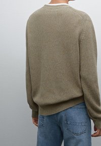 Person, der einen beigen Strickpullover und helle Jeans trägt, von hinten vor einem einfarbigen grauen Hintergrund gezeigt.