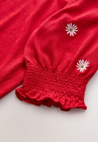 Red knit sweater featuring a smocked cuff with a frilled edge. White daisy embroidery with a brown center on each sleeve.
